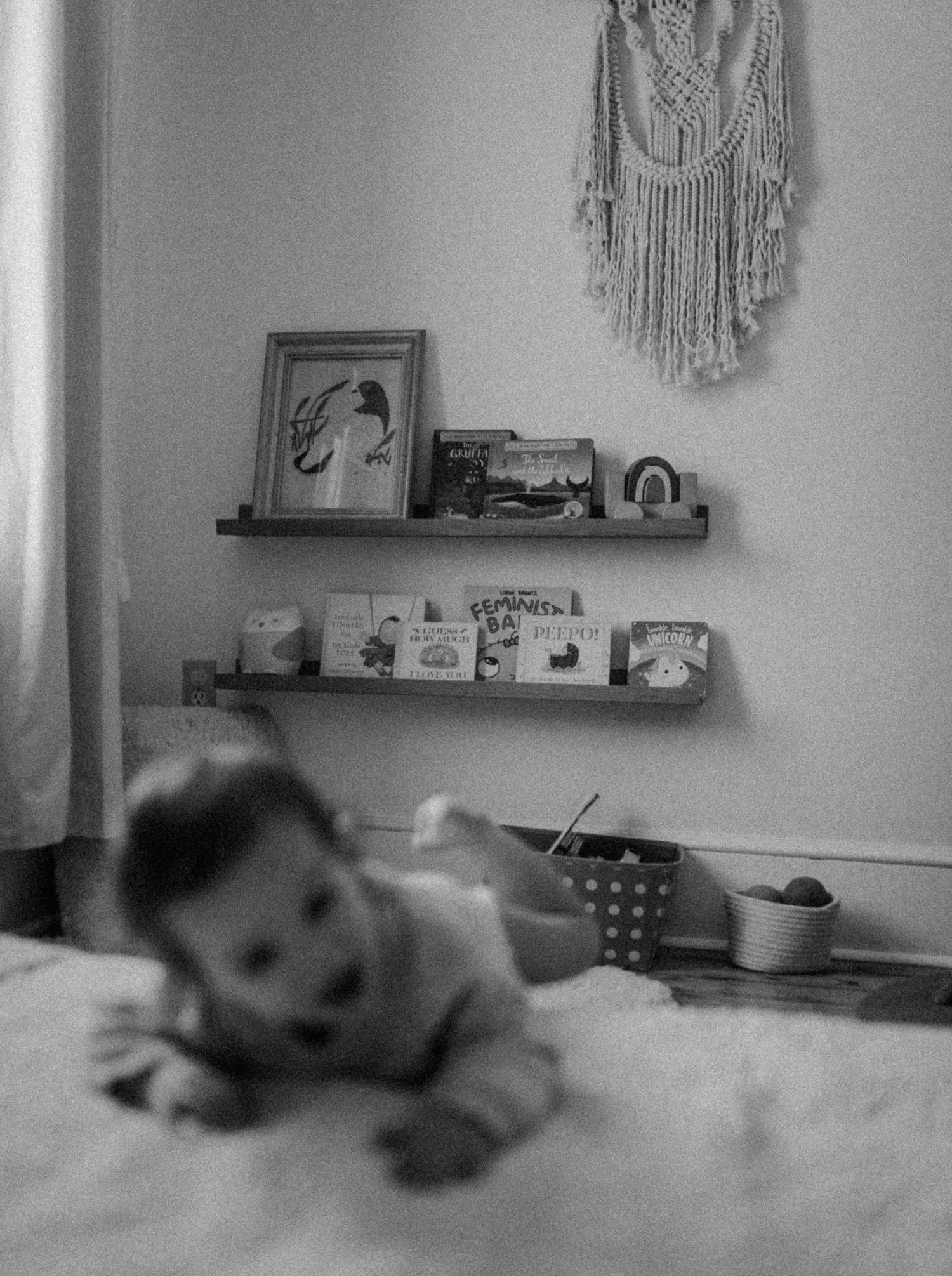 Black and white photo of a nursery corner with macrame wall art and floating shelves displaying books.