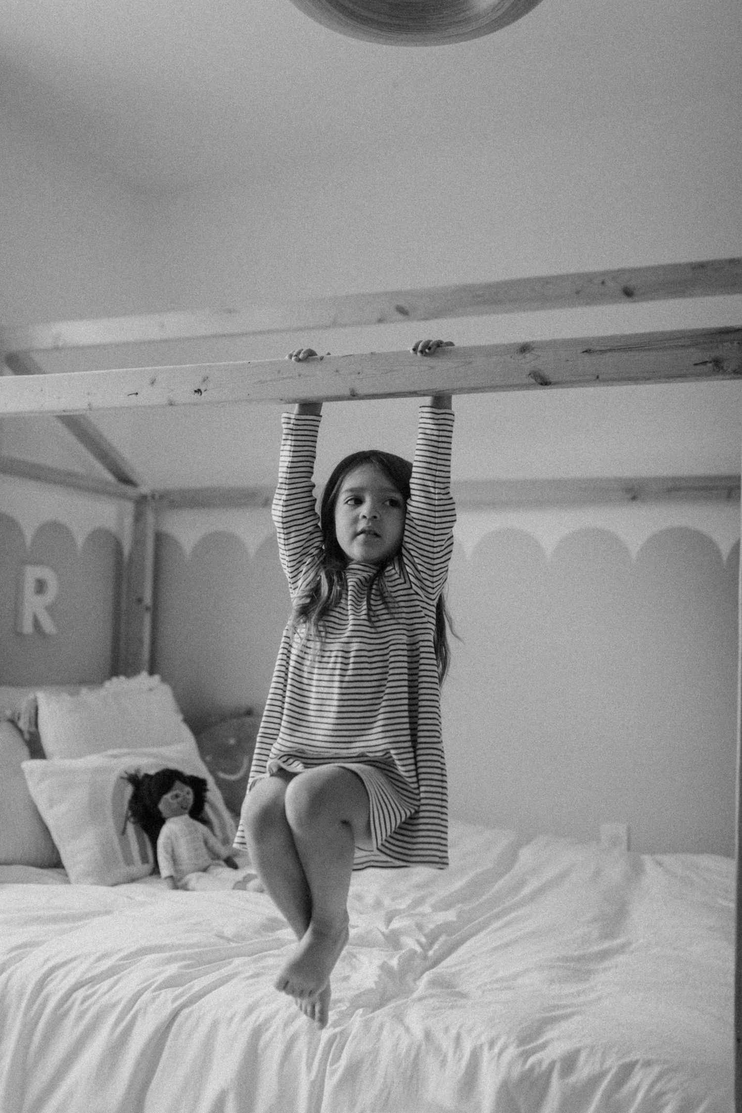 A child in striped clothing hanging from wooden beams above a white bed.