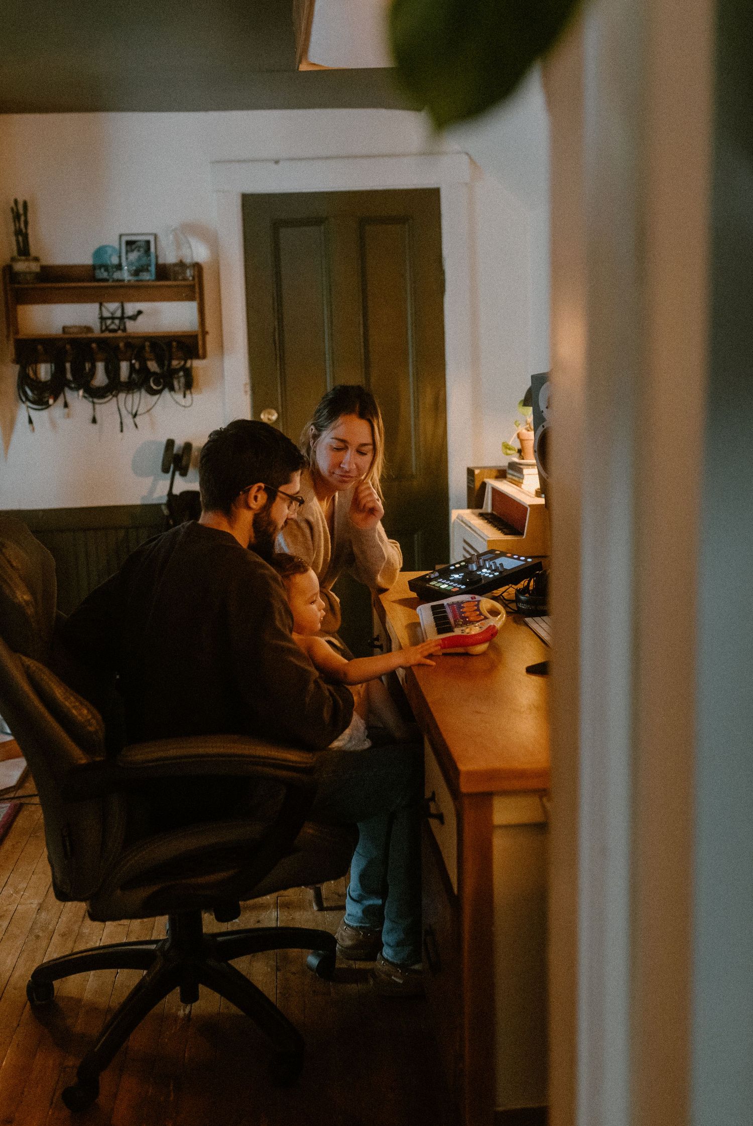 Small family gathered around a desk in cozy home office with warm lighting and rustic decor on wall.