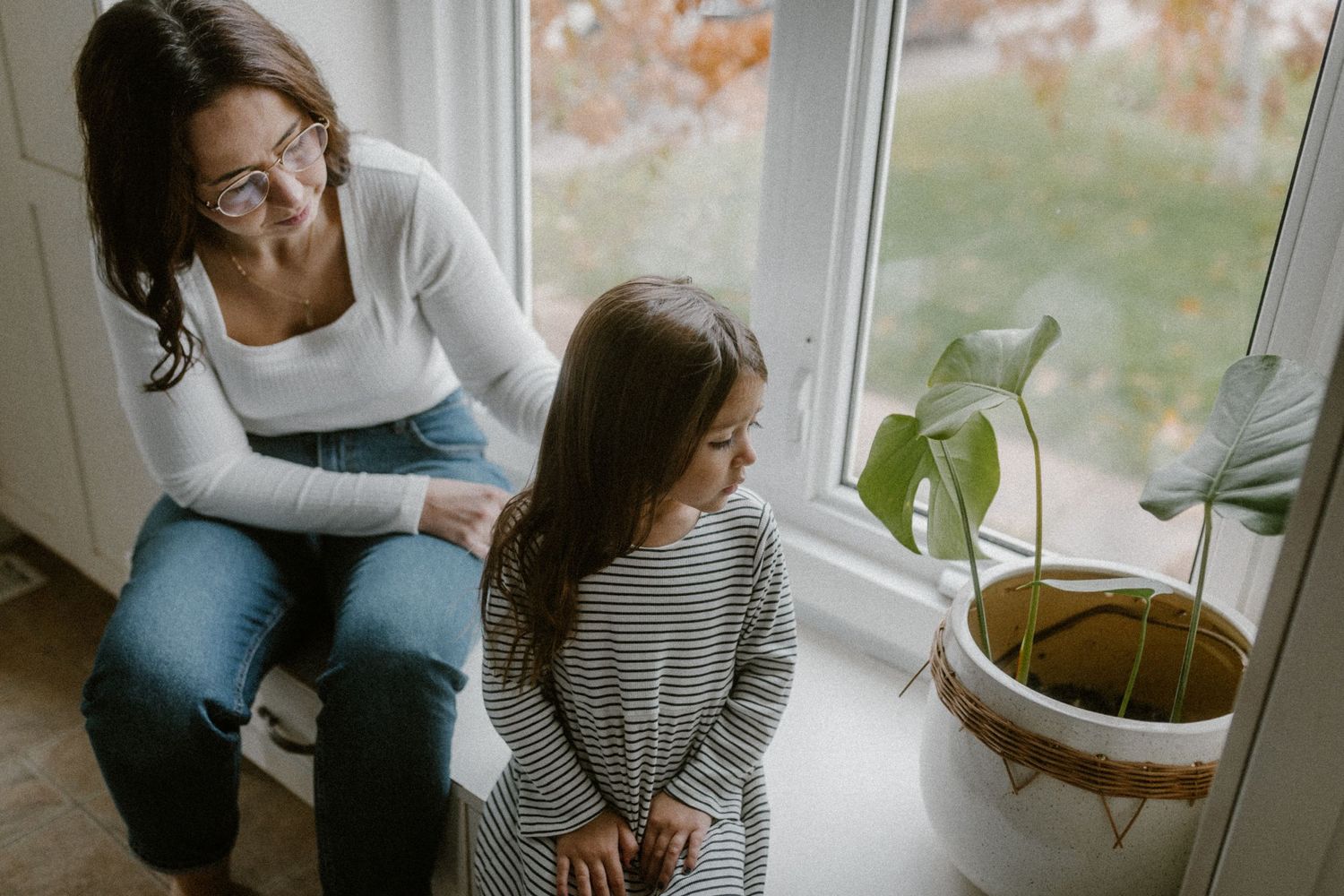 Mom and daughter sit by a sunny window with houseplants having an intimate conversation.