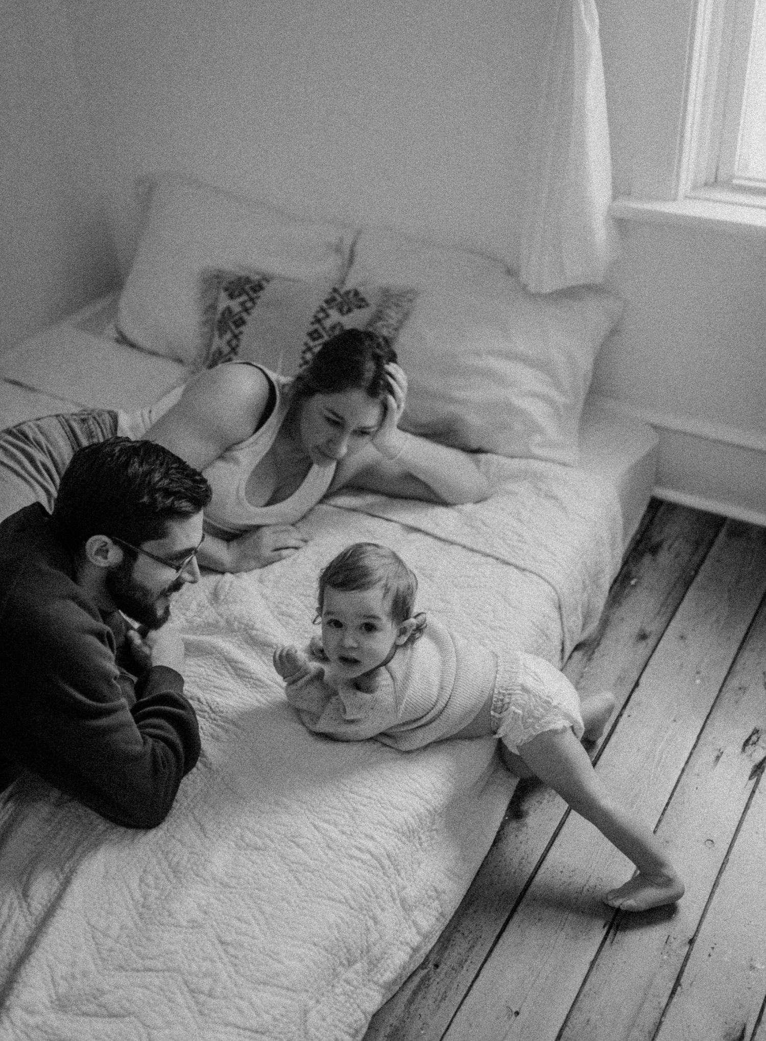 Family moment captured in black and white as they relax together on a mattress with wooden flooring.