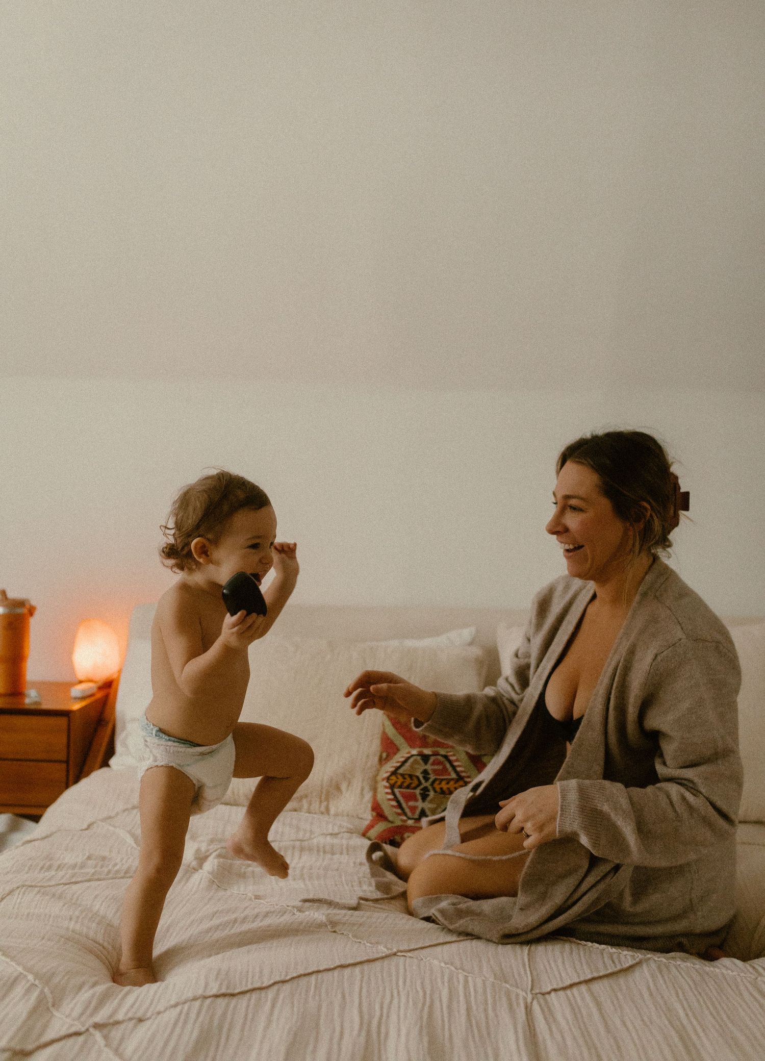 A candid moment in a warmly lit bedroom shows a playful scene between parent and toddler on a white bed.