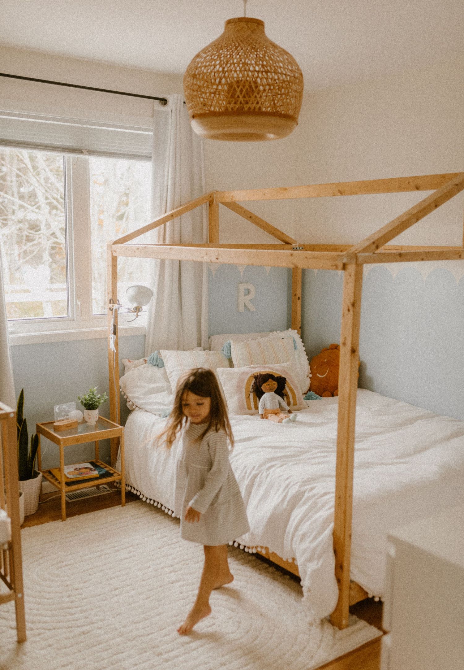 A cozy children's bedroom with a wooden house frame bed, woven pendant lamp, and white bedding in natural lighting.