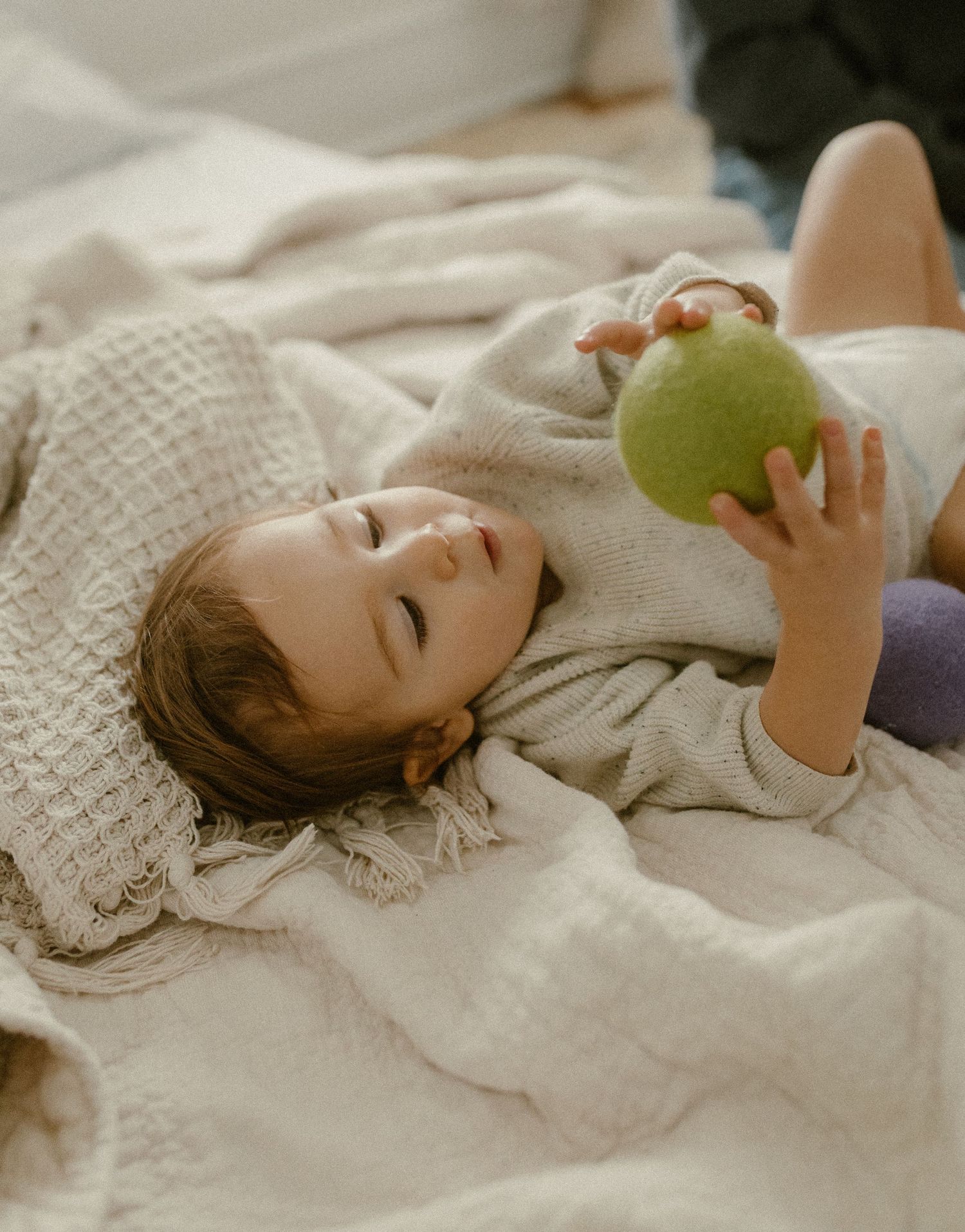 Child lying on white bedding holding a green ball during playtime.