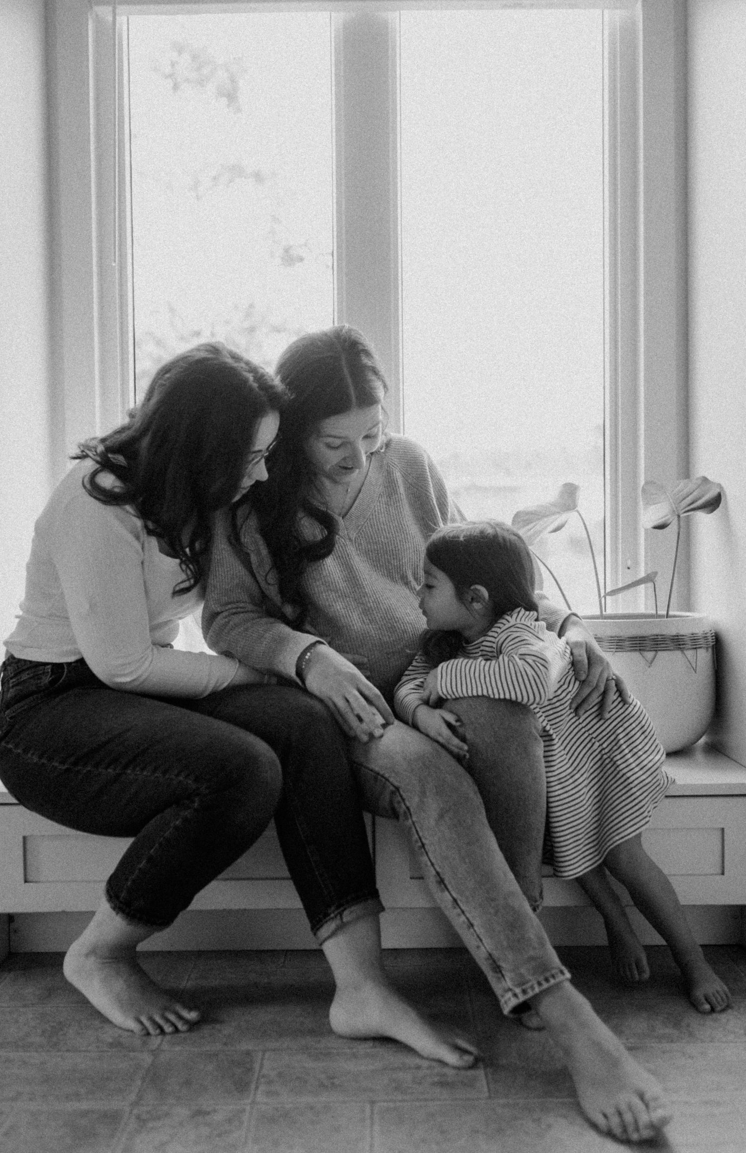 A heartwarming black and white photograph shows a tender family moment by a window, with everyone sitting barefoot together.