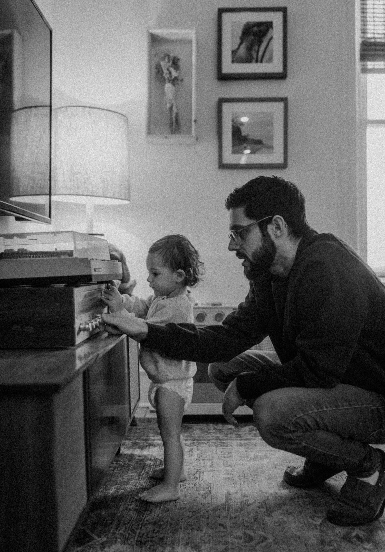 Black and white photo of family interaction near a vintage piano with lamp and framed artwork.
