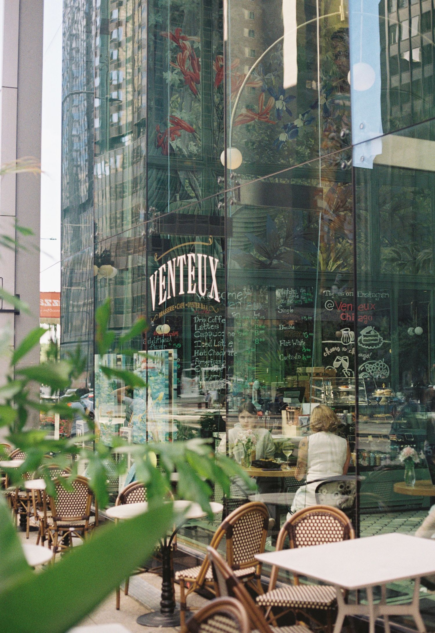 Modern cafe interior with glass walls reflecting city buildings and wooden bistro chairs arranged on outdoor terrace.