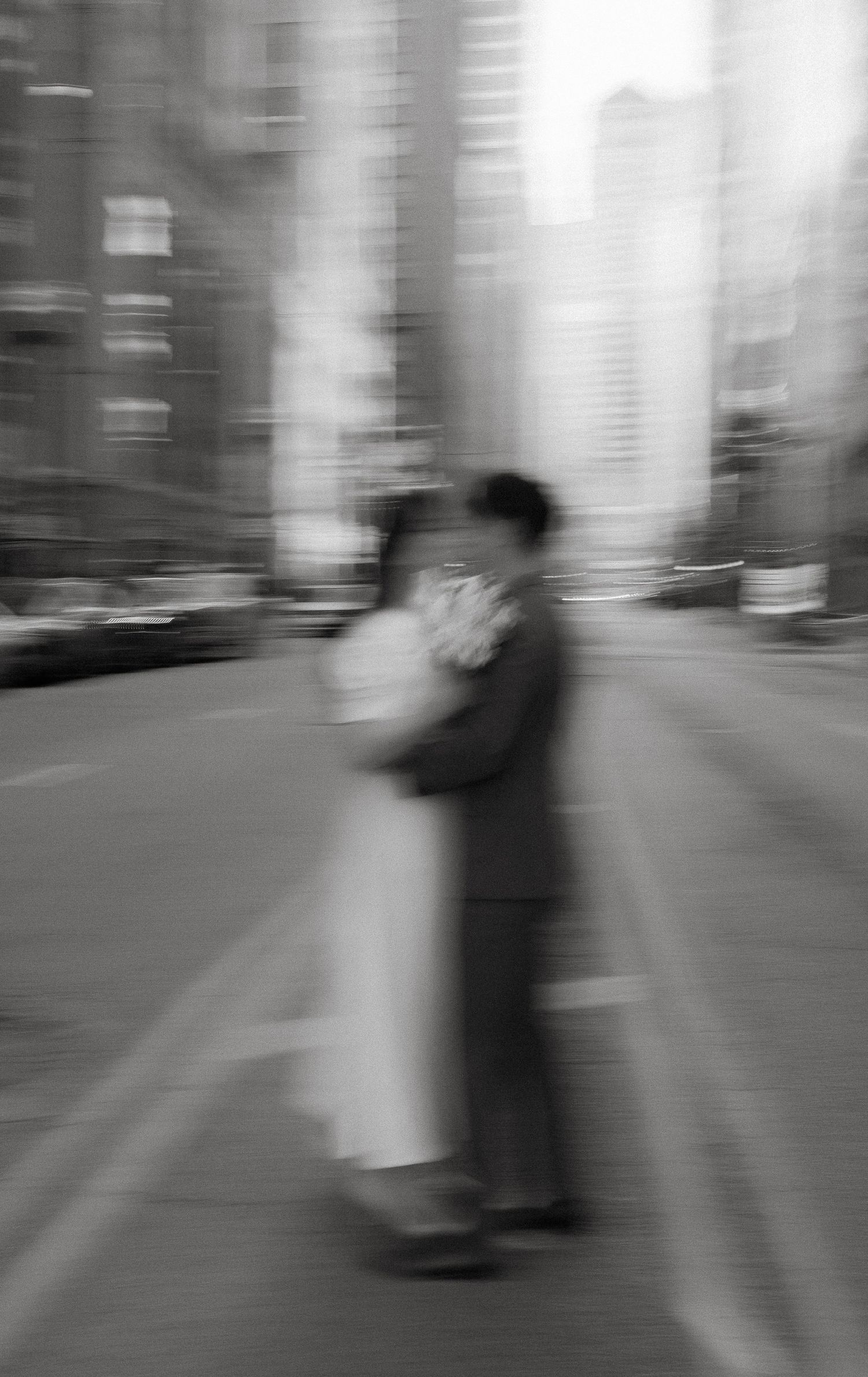 A blurred black and white artistic photograph of two people embracing on an urban street with tall buildings in background.