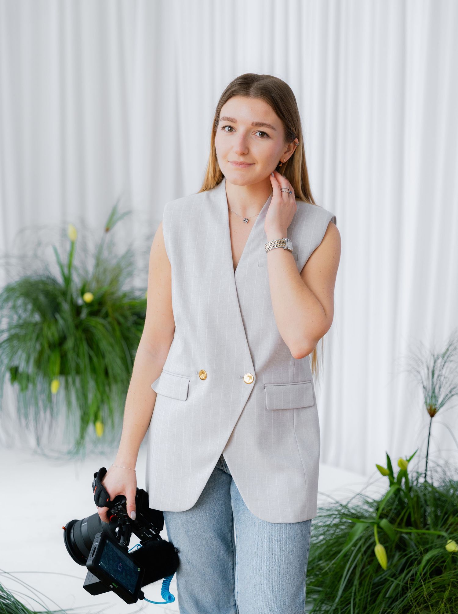 Woman in gray vest and jeans poses with black cat in modern room with white curtains and green plants.