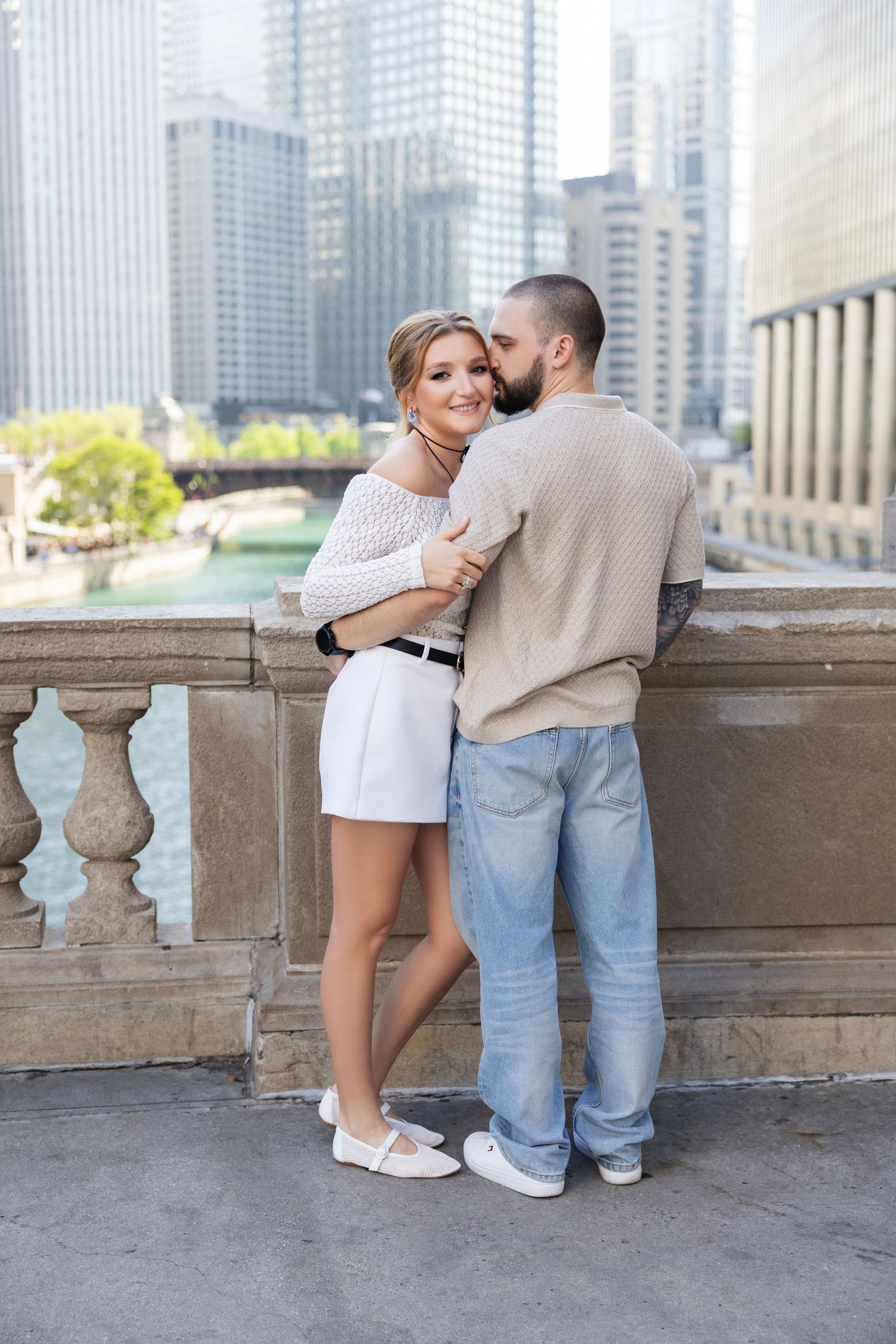 A couple shares a romantic moment while leaning against a stone balcony with a city skyline in the background.