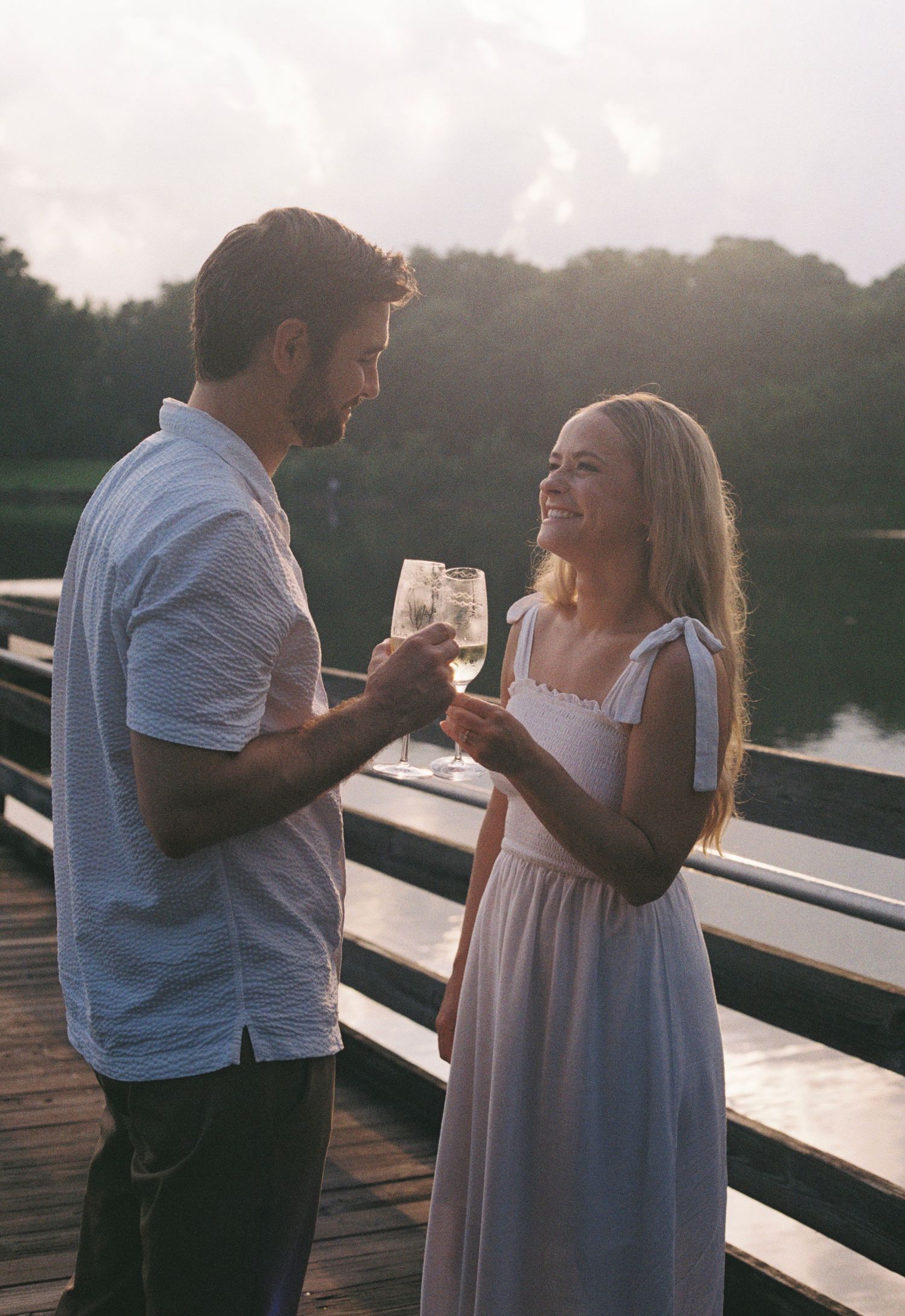 Couple shares romantic moments at sunset on a lakeside dock while enjoying drinks together.