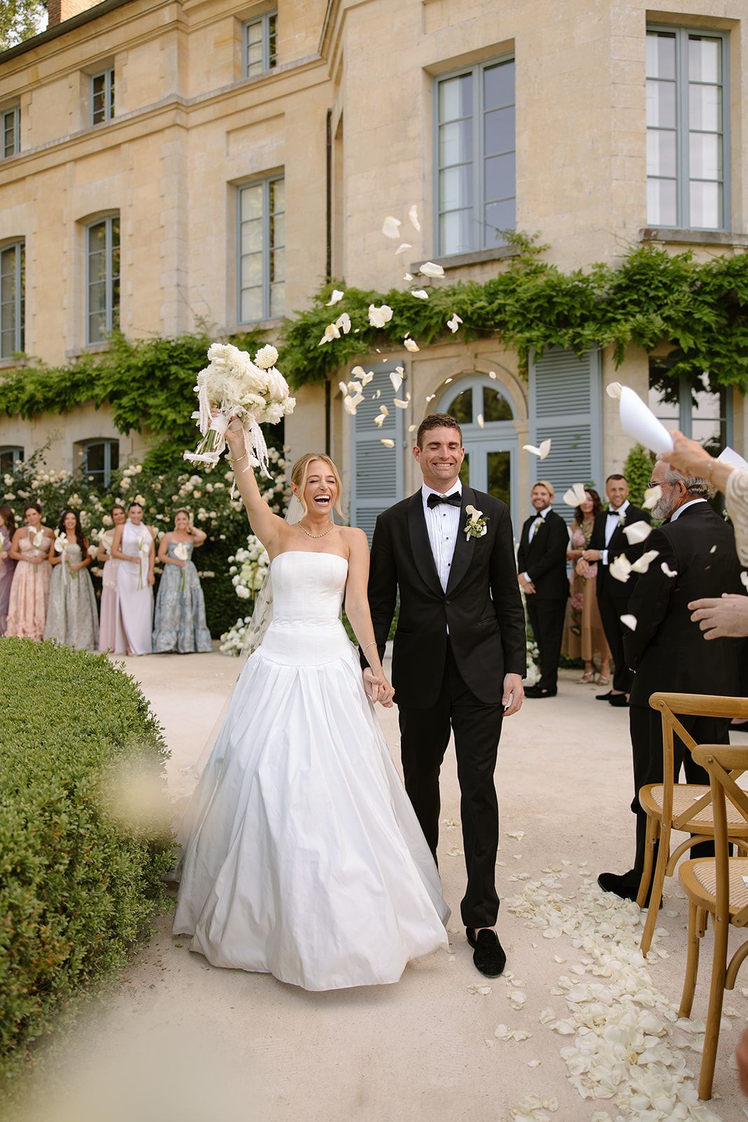 Bride in strapless white ball gown and groom in black tuxedo walk joyfully through petal toss at elegant French chateau wedding.