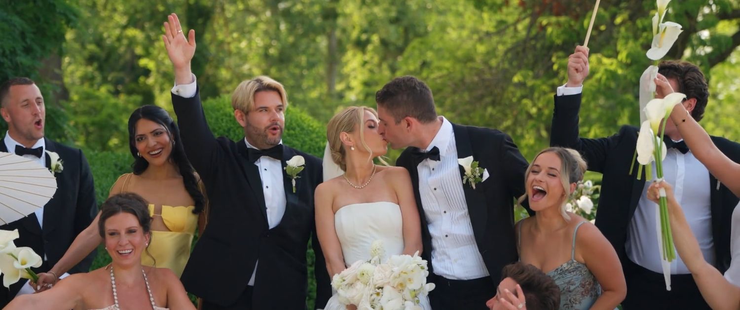 Wedding party celebrating outdoors, bride and groom kissing surrounded by cheering bridesmaids and groomsmen in formal attire.