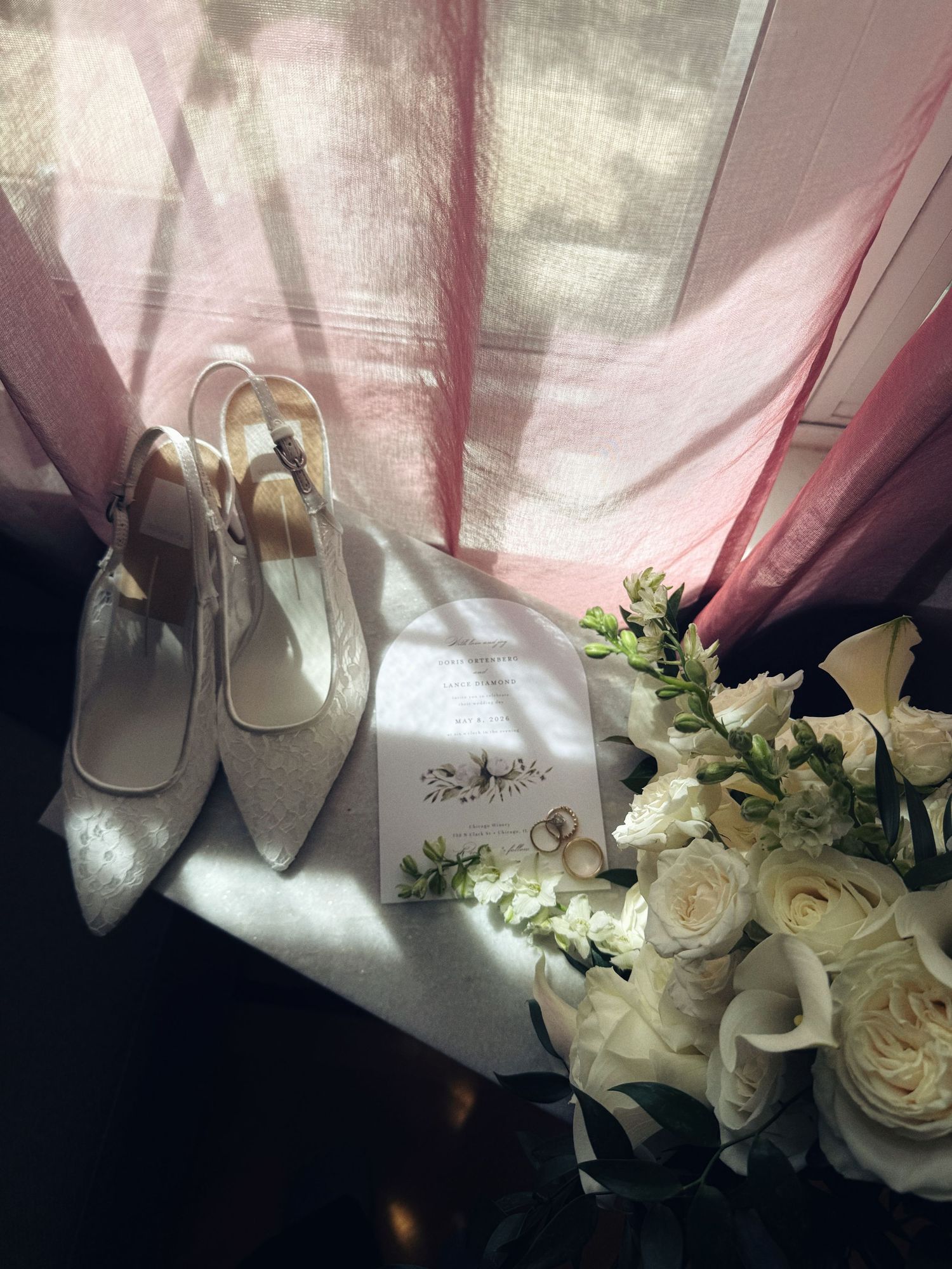 Elegant bridal flat lay with white lace slingback heels, gold wedding rings, floral invitation, and white rose bouquet by a window.