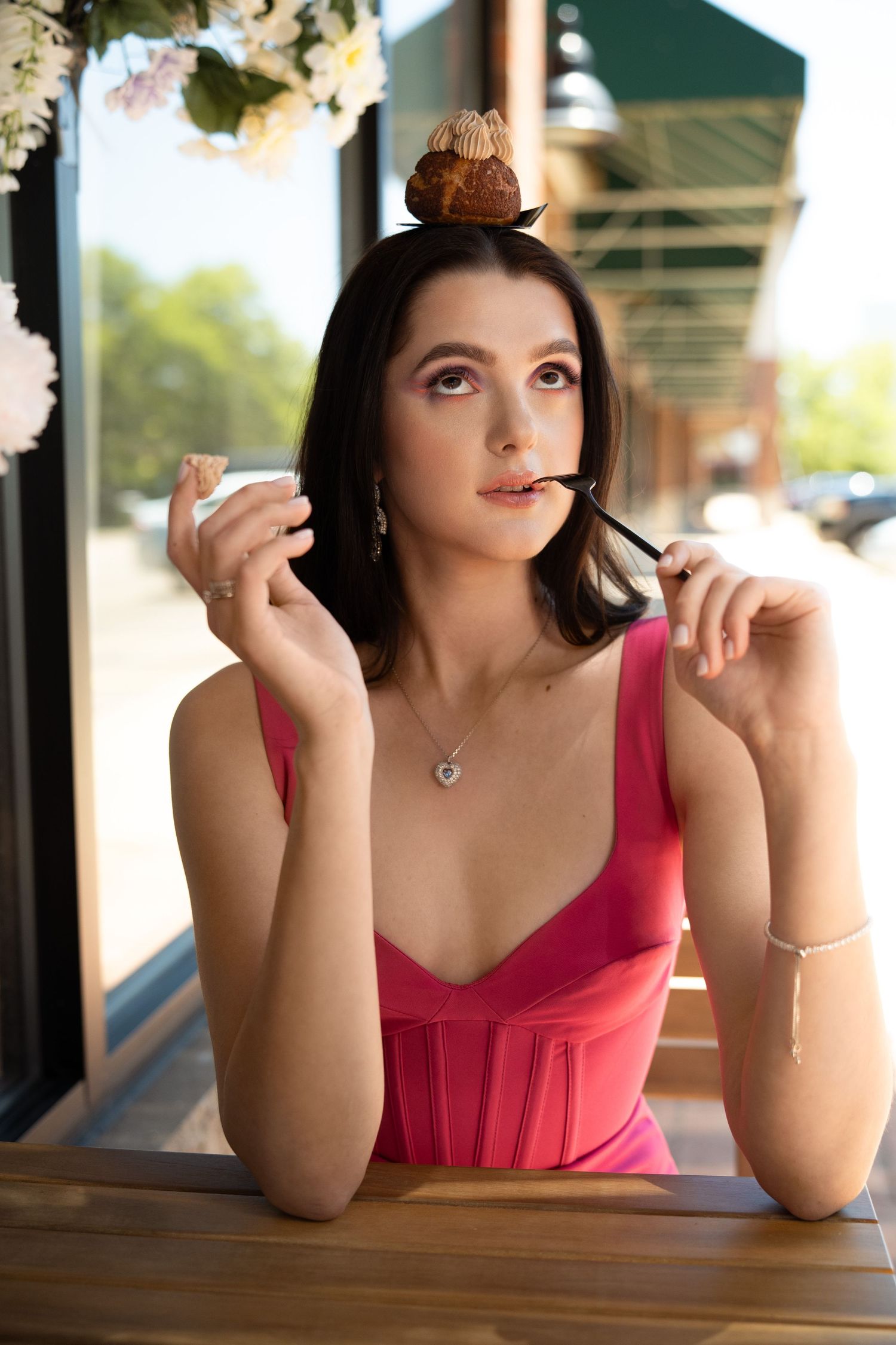Woman in a pink corset top sitting at an outdoor table with a small brown pastry balanced on her head.