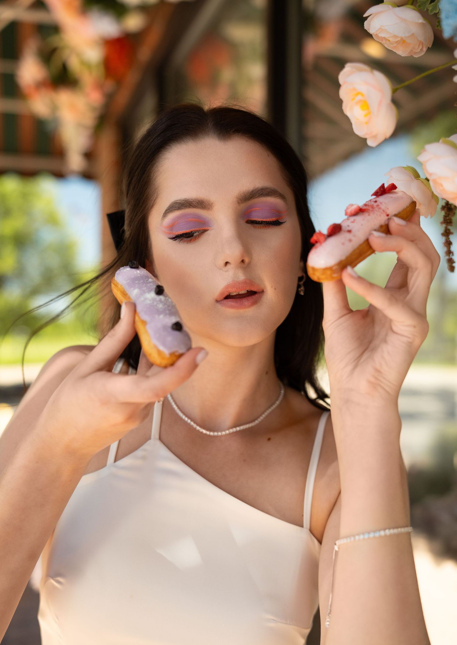 Woman with pink eyeshadow and closed eyes holding colorful macarons near blooming flowers.