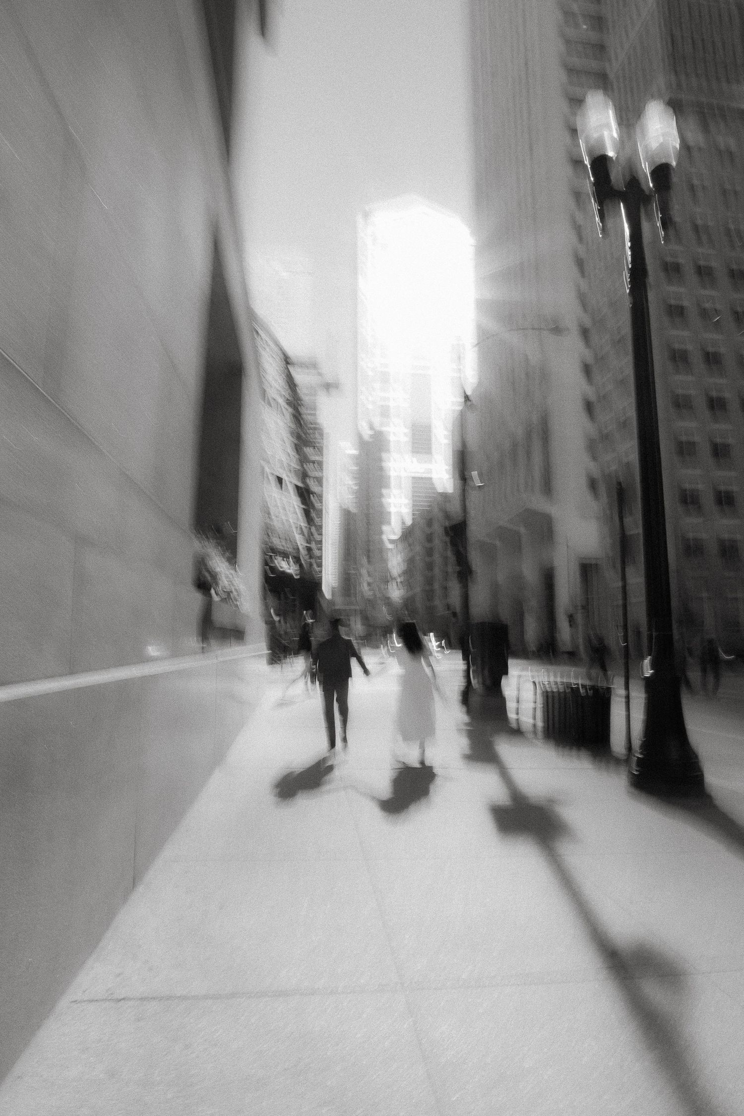 A black and white blurred cityscape with shadowy figures walking on a sidewalk beneath street lamps and tall buildings.