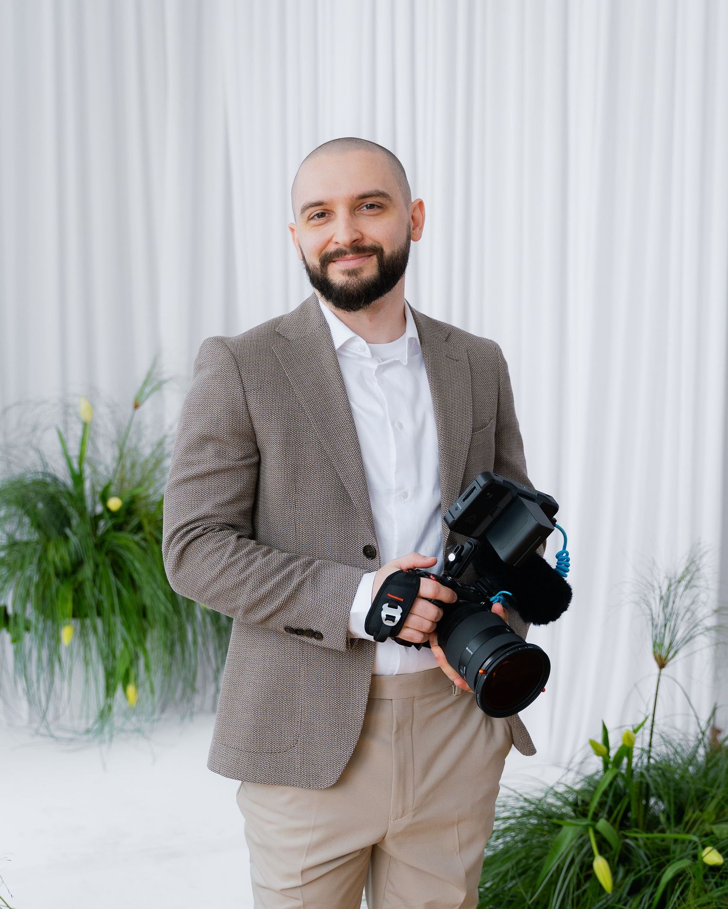 Professional photographer in beige suit holding camera against white backdrop with green plants.