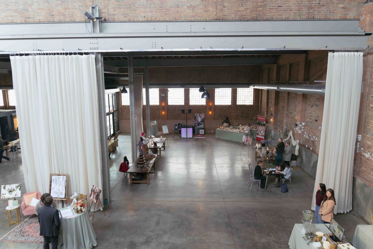 Large industrial event space with white curtain dividers and brick walls hosts vendor tables. At Fairlie, Chicago