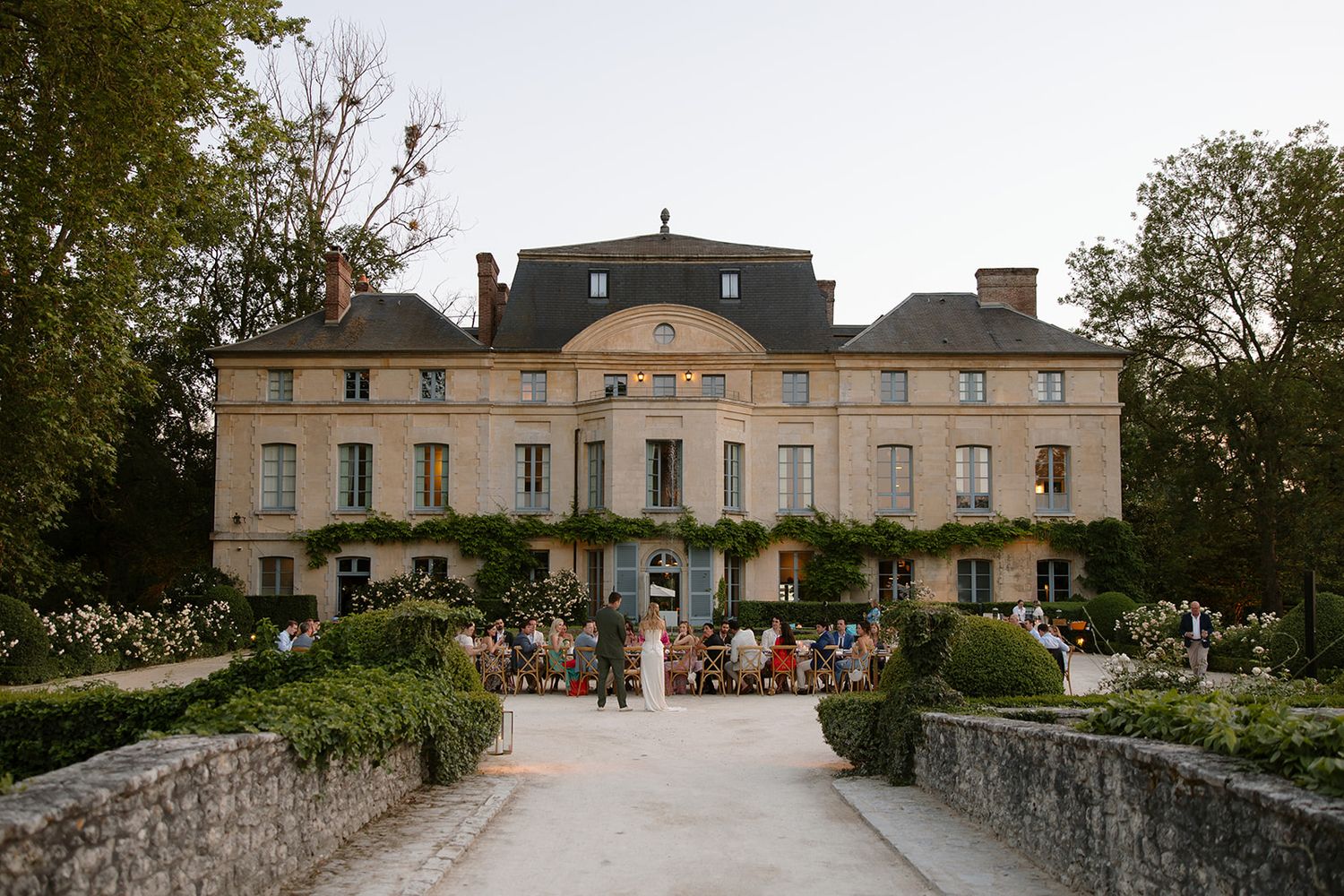 Elegant French chateau wedding venue surrounded by manicured gardens and trees at dusk with guests gathered outside.