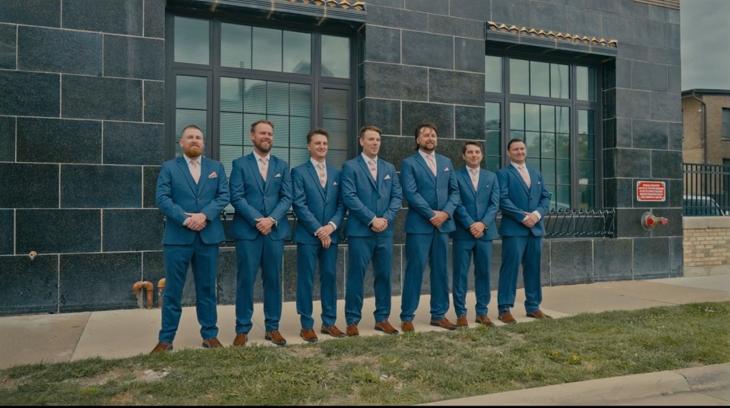 A wedding party of six groomsmen standing in a line wearing matching navy blue suits with tan shoes outside a building.