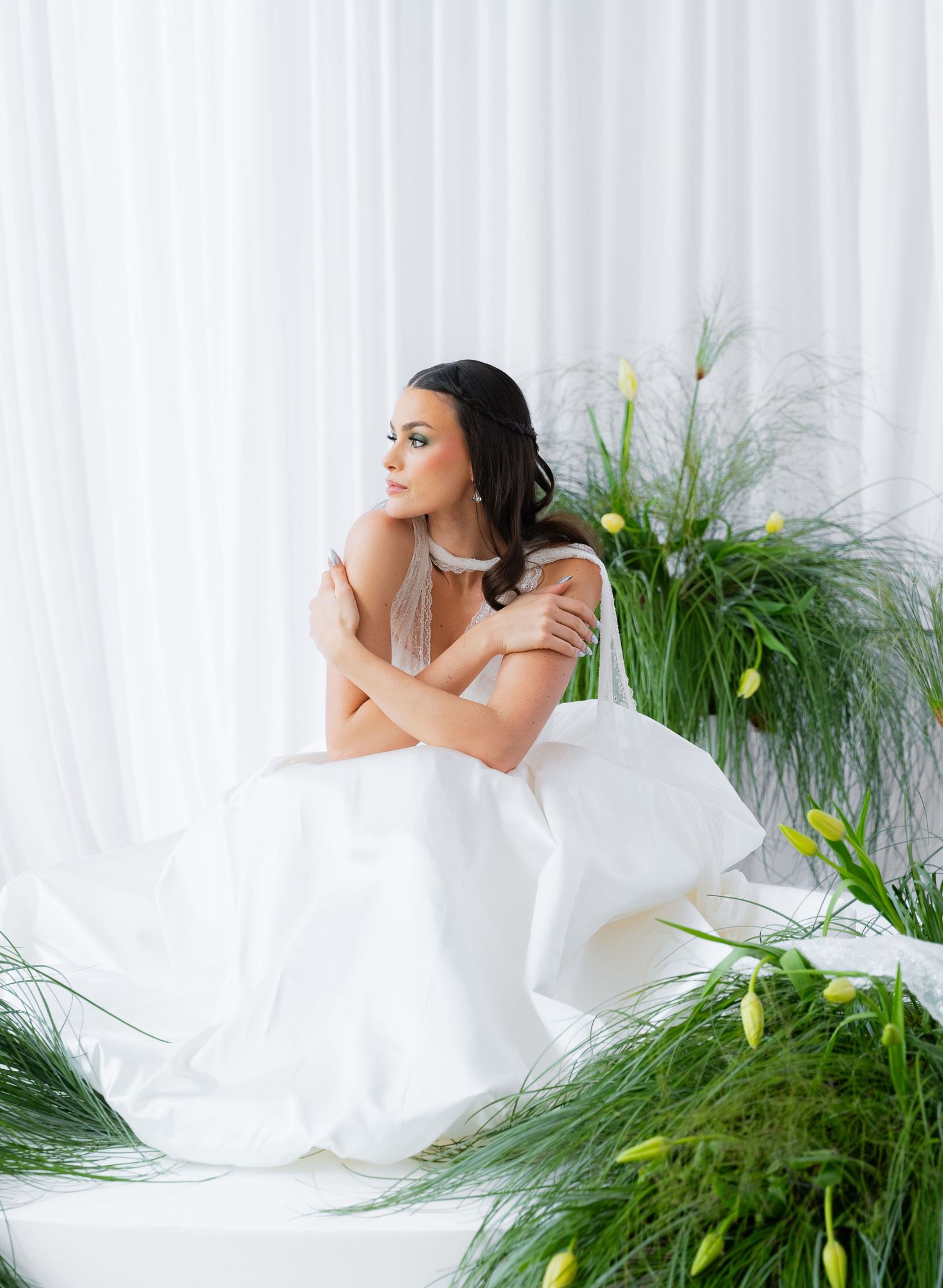 Elegant bridal portrait with flowing white gown against sheer curtains and decorative greenery arrangements.