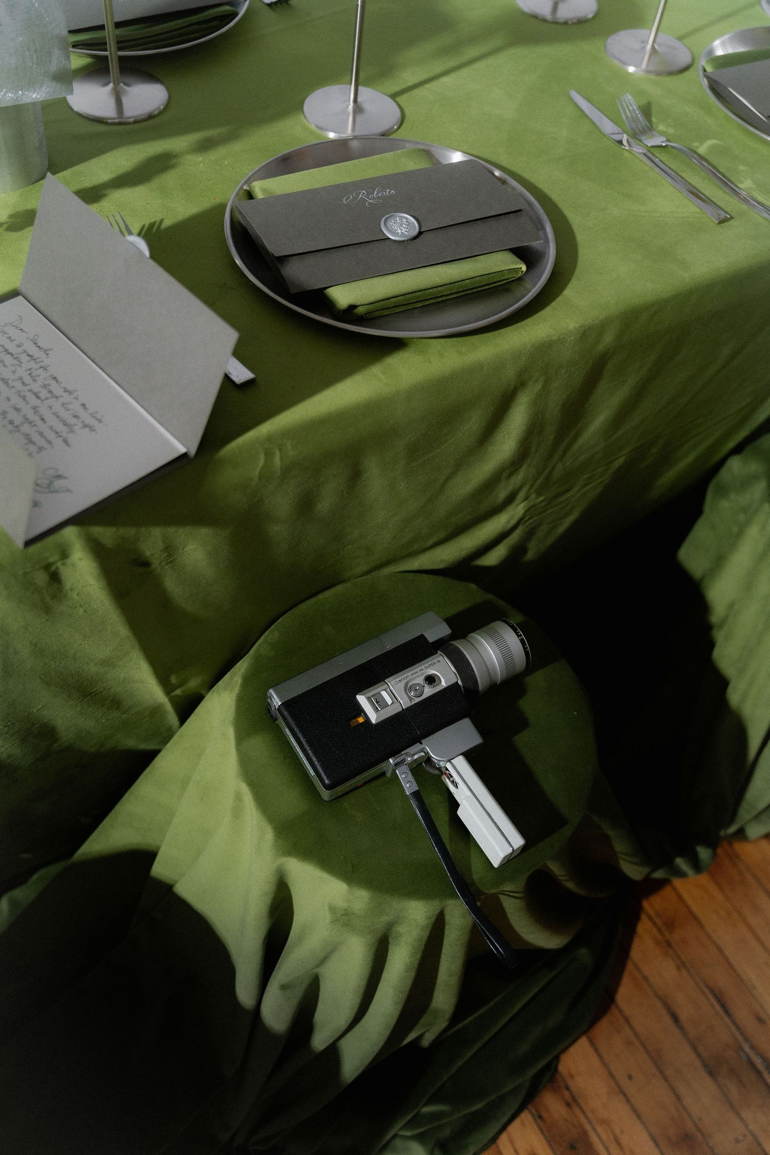 A green tablecloth being laid on a dining table with dark plates and silverware visible.