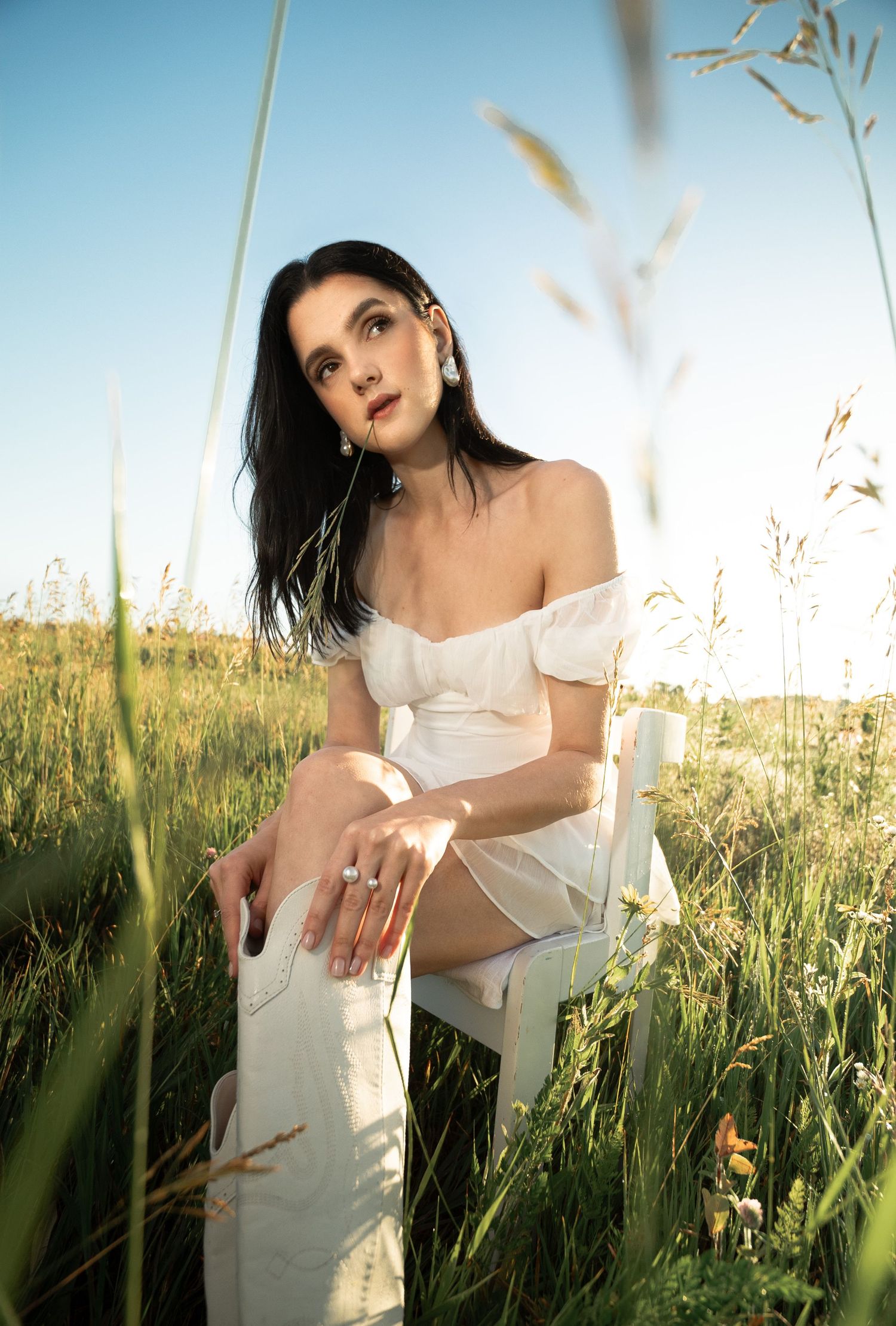 Woman in white dress sitting on chair in sunlit field of tall grass during golden hour.