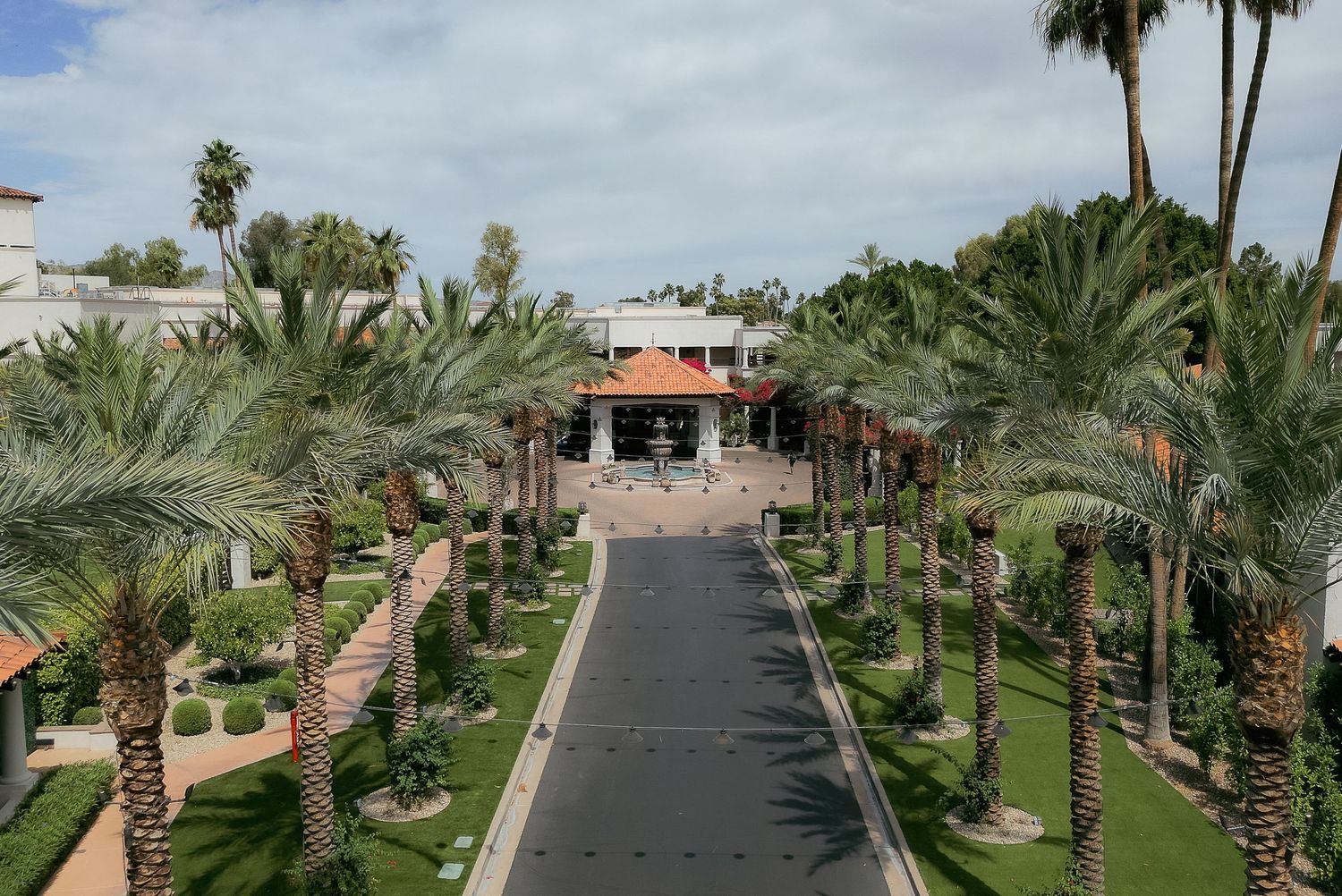 Palm tree-lined driveway leading to Spanish-style entrance of resort building under blue sky.
