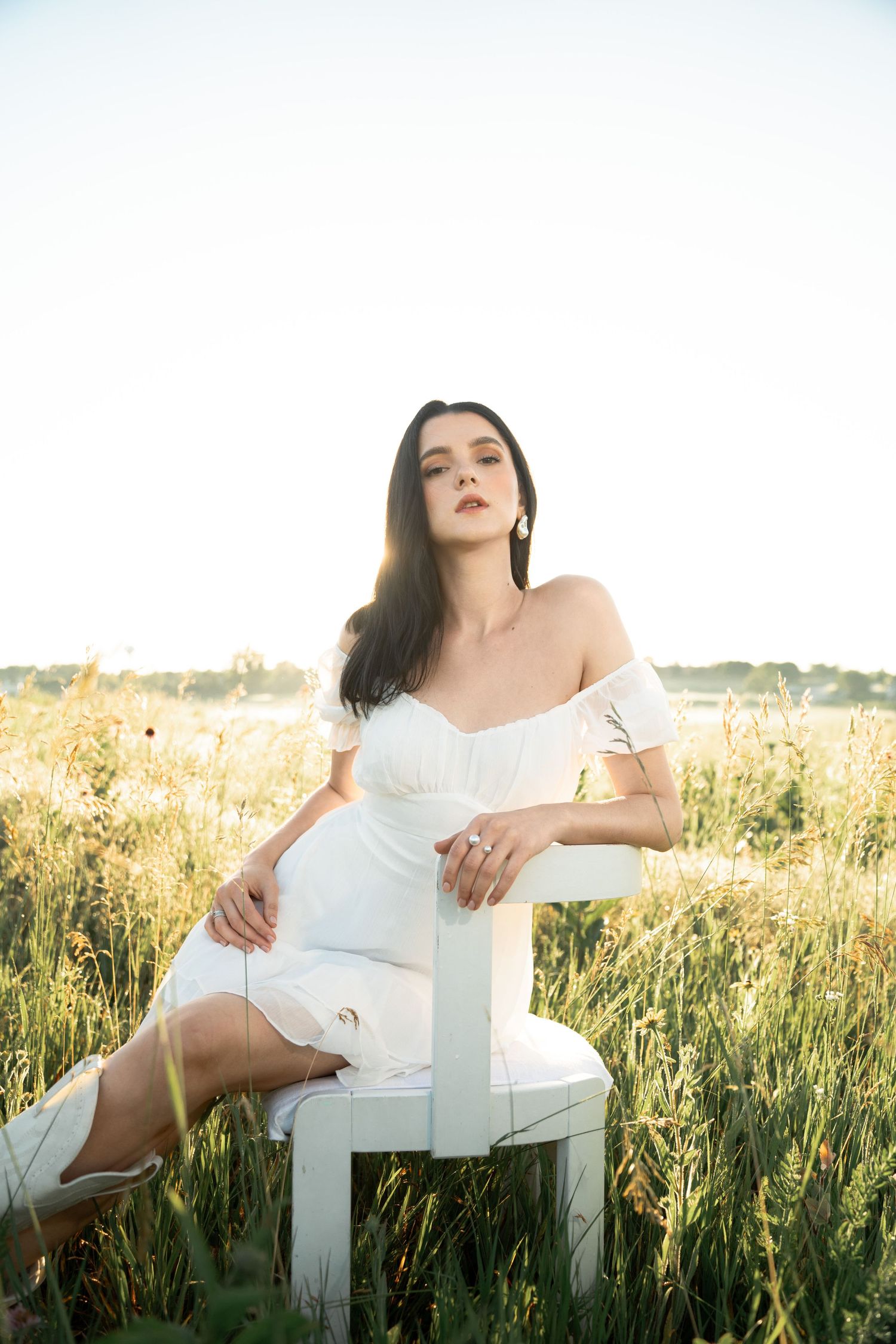 Woman in white dress sitting elegantly on white chair surrounded by summer meadow grass.
