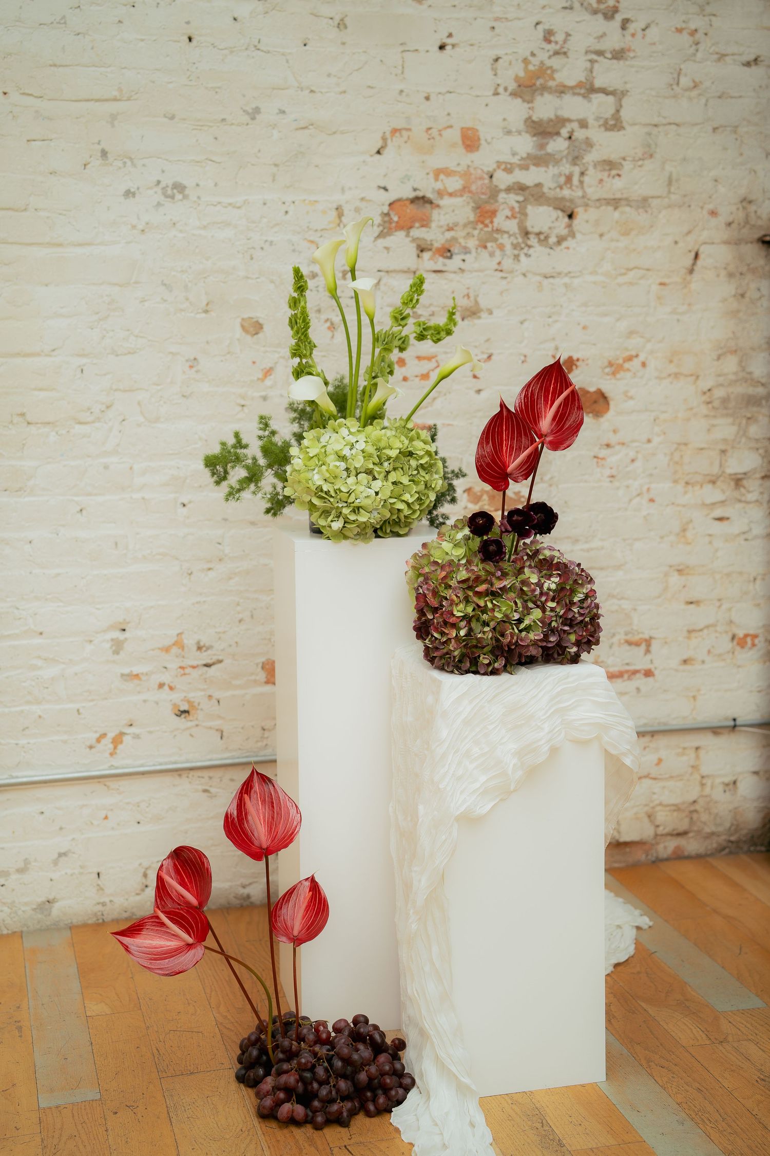 Modern floral arrangement with red anthurium flowers and green hydrangeas in white vases against a rustic wall background.