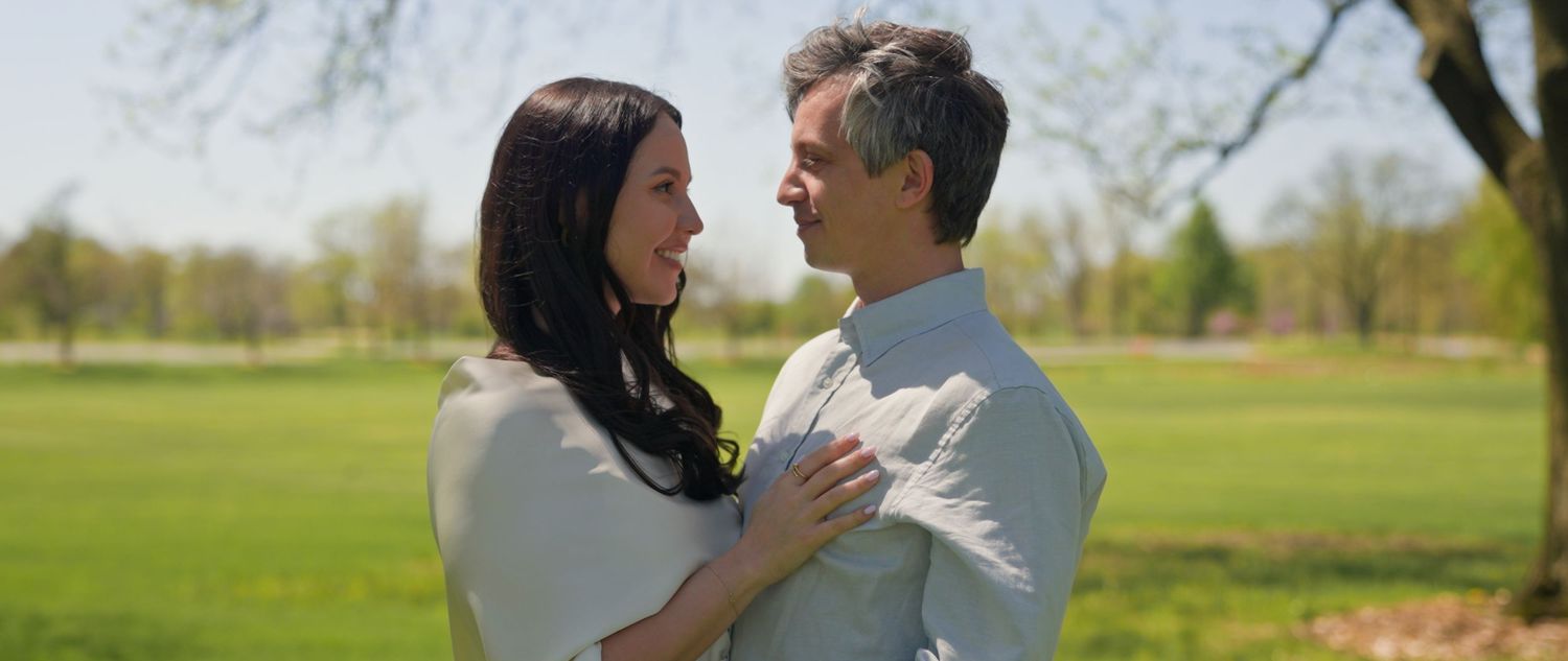 A couple smiles at each other in a sunny park, the woman placing her hands on the mans chest affectionately.