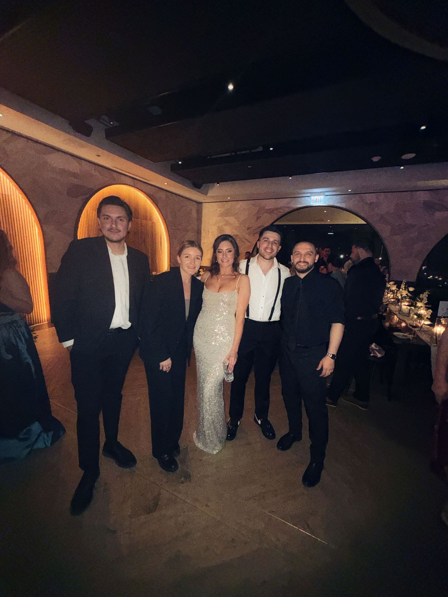 Five elegantly dressed people posing together at a formal evening event in a dimly lit venue with arched decor.