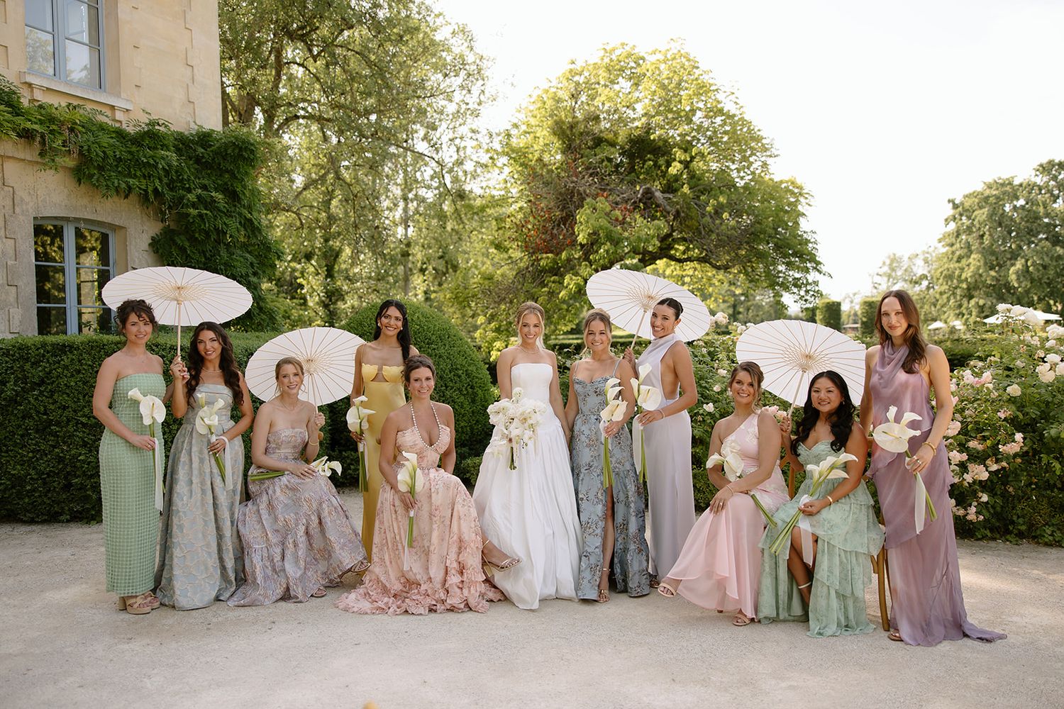 Bride in white gown with bridesmaids in pastel dresses holding parasols posing outdoors at elegant garden wedding venue.