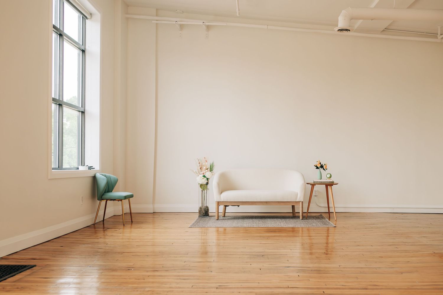 Minimalist studio space with hardwood floors featuring a teal chair, cream-colored bench, and side tables near a window.