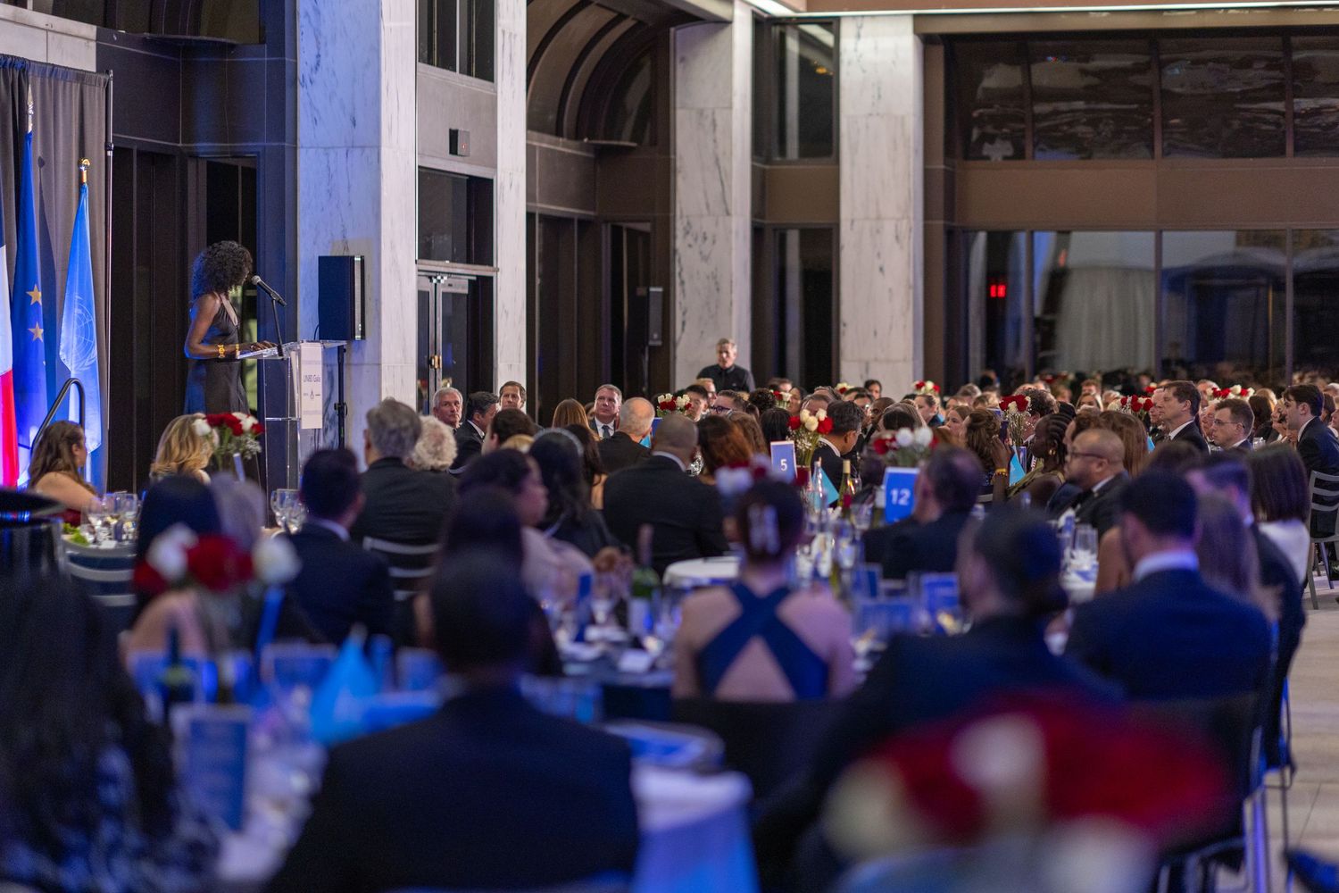 Business conference attendees seated in rows at a large venue with blue lighting and presentation screens.