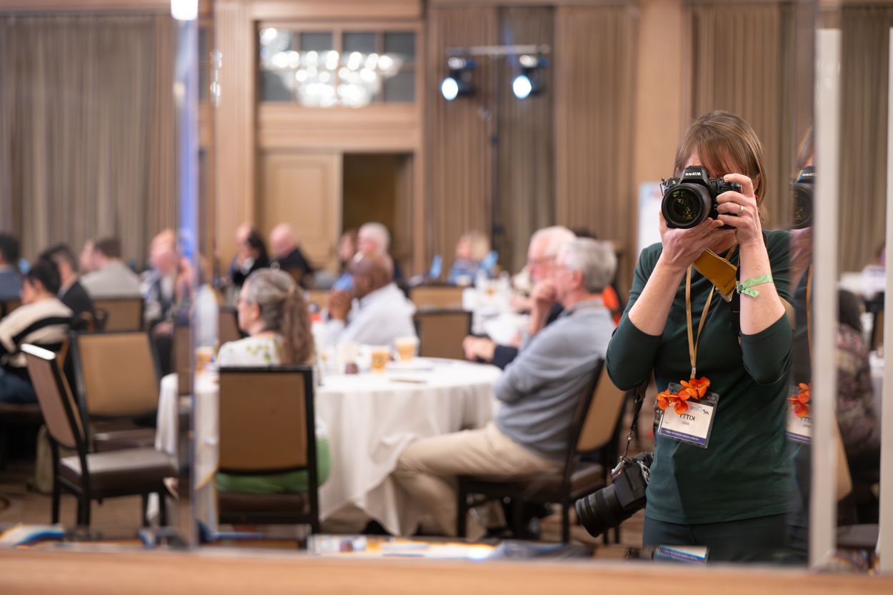 Woman photographer taking a self-portrait in a mirror at a formal banquet hall event with guests seated at round tables.