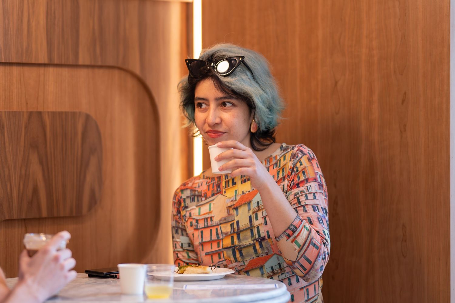 Woman with blue hair and sunglasses on head sits at a table eating in a modern wood-paneled restaurant.