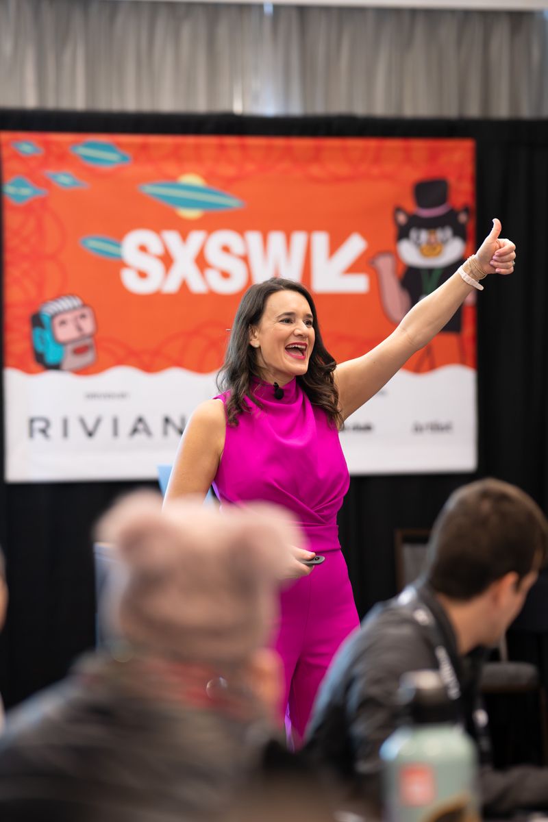 Woman in pink dress giving thumbs up at SXSW event with Rivian sponsored backdrop.