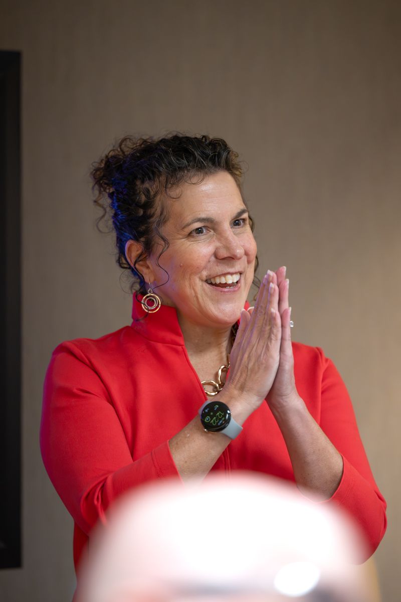 A smiling woman in a red blazer claps her hands together, wearing colorful earrings and a smartwatch.
