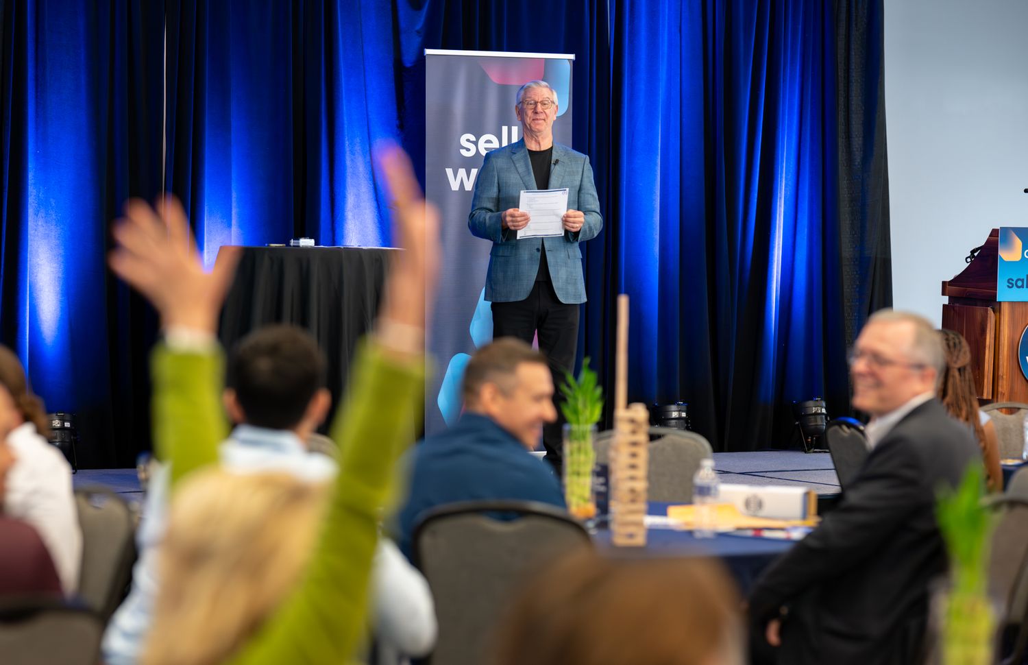 Speaker addresses engaged audience with raised hands at a professional conference or seminar event.