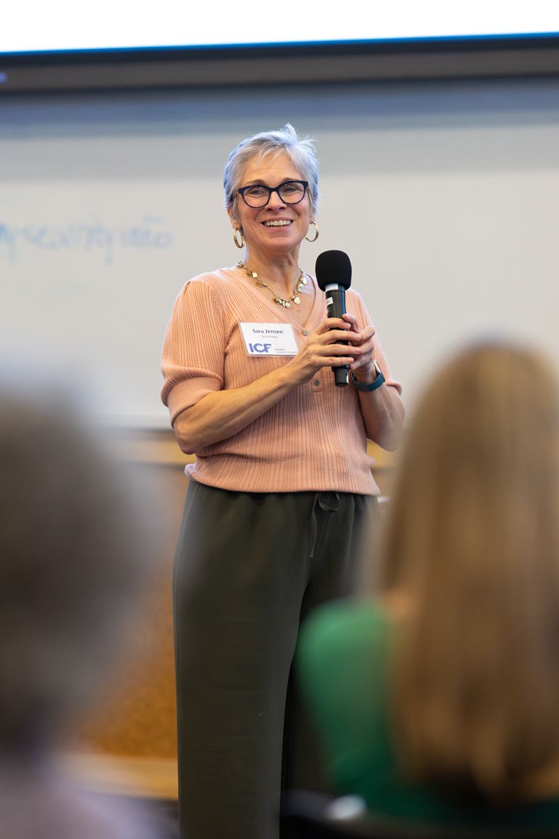 A person with short gray hair and glasses speaks into a microphone while wearing a peach colored top and dark skirt.