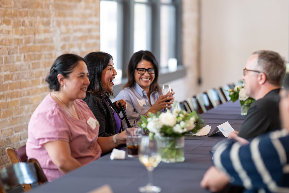 Three women and a man laugh together at a long conference table with wine glasses and floral centerpieces.