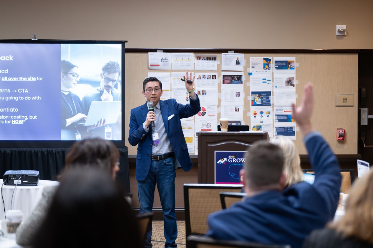 Speaker presenting at a conference with a microphone, gesturing to audience with a PowerPoint slide displayed behind him.
