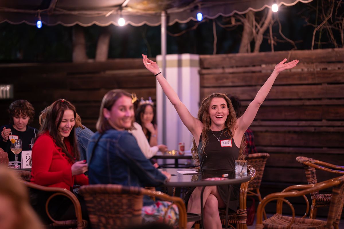 Woman raises arms in celebration at outdoor evening gathering with friends seated at tables under string lights.