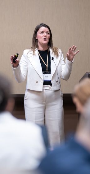 Woman in white suit speaks at conference, gesturing expressively while holding a clicker and wearing a name badge.