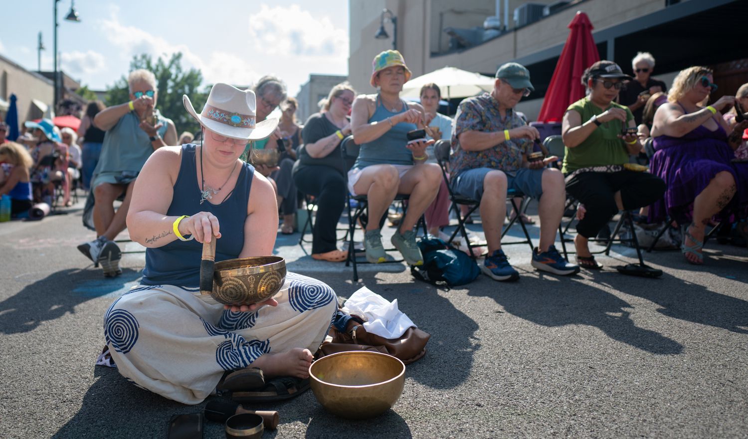 People playing Tibetan singing bowls outdoors at a sunny street festival, seated in chairs and on the ground.