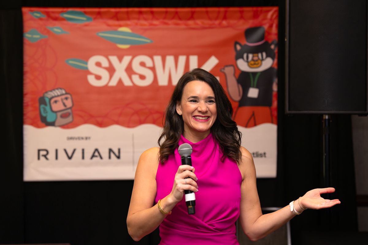 Woman in pink dress speaking at SXSW event on stage with microphone, Rivian sponsored backdrop.