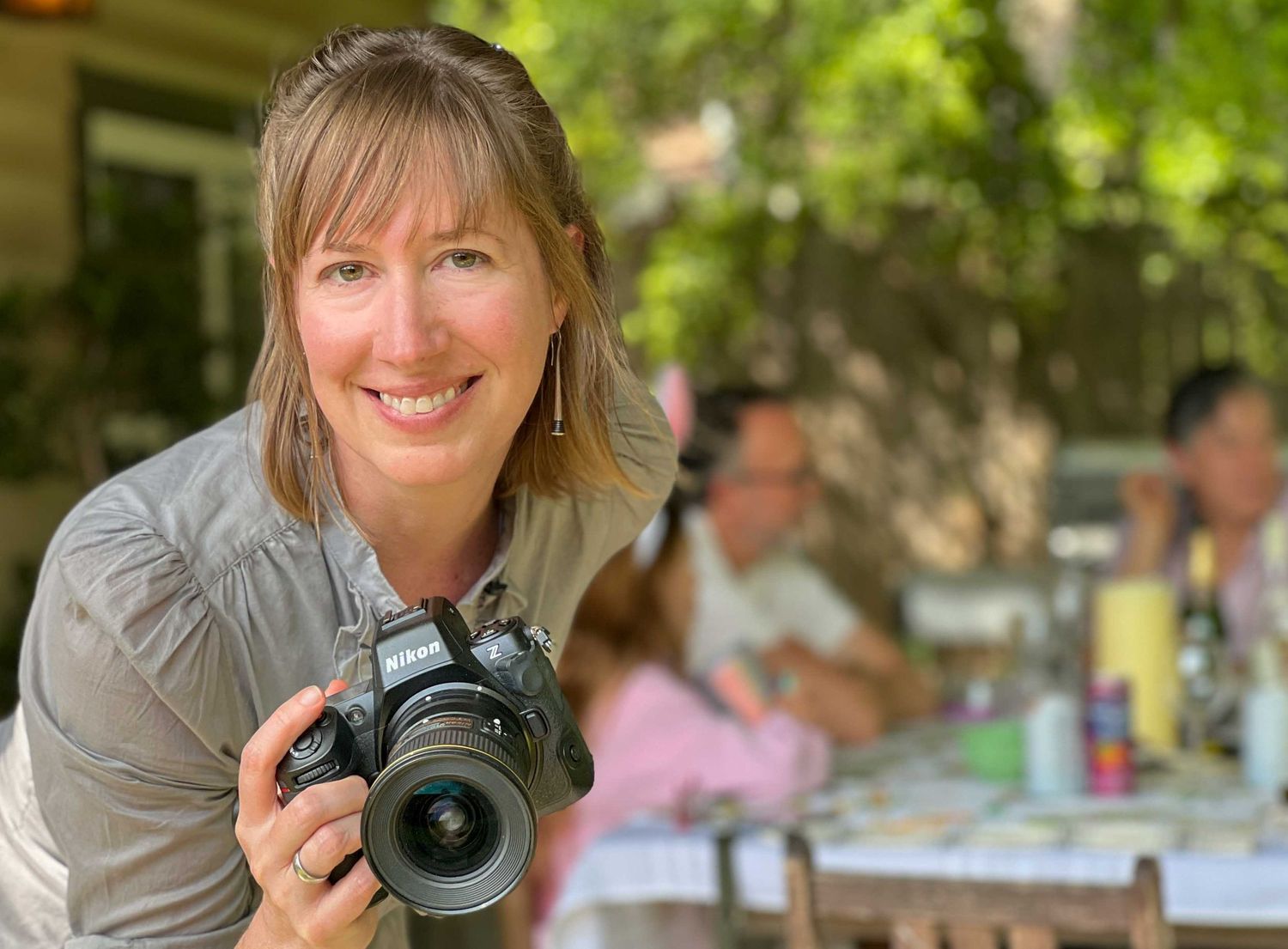 A photographer in a gray shirt holds a professional camera while smiling outdoors in natural lighting.