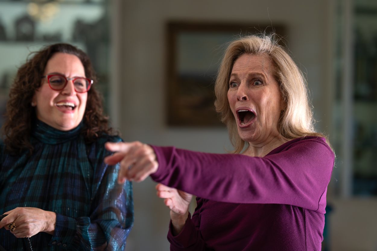 Two women laughing and pointing excitedly indoors, one in teal plaid, one in purple top.